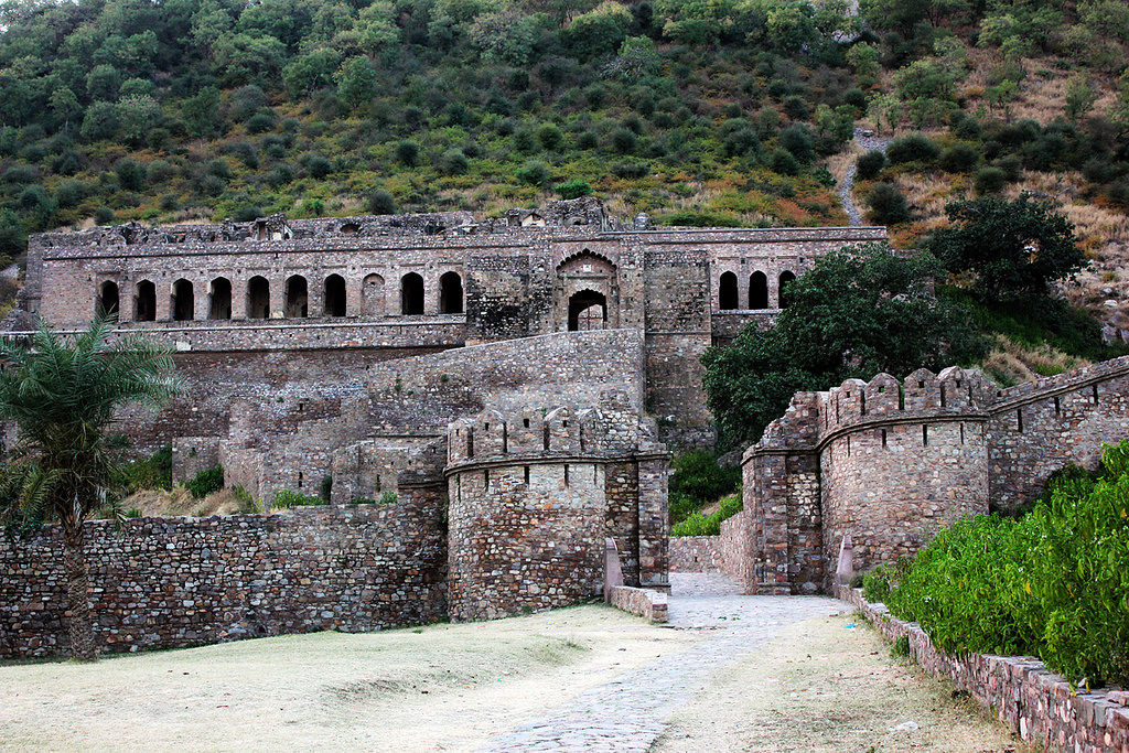 Ancient stone fortress ruins with arched windows and battlement walls set against a green forested hillside, muted grey and ochre tones