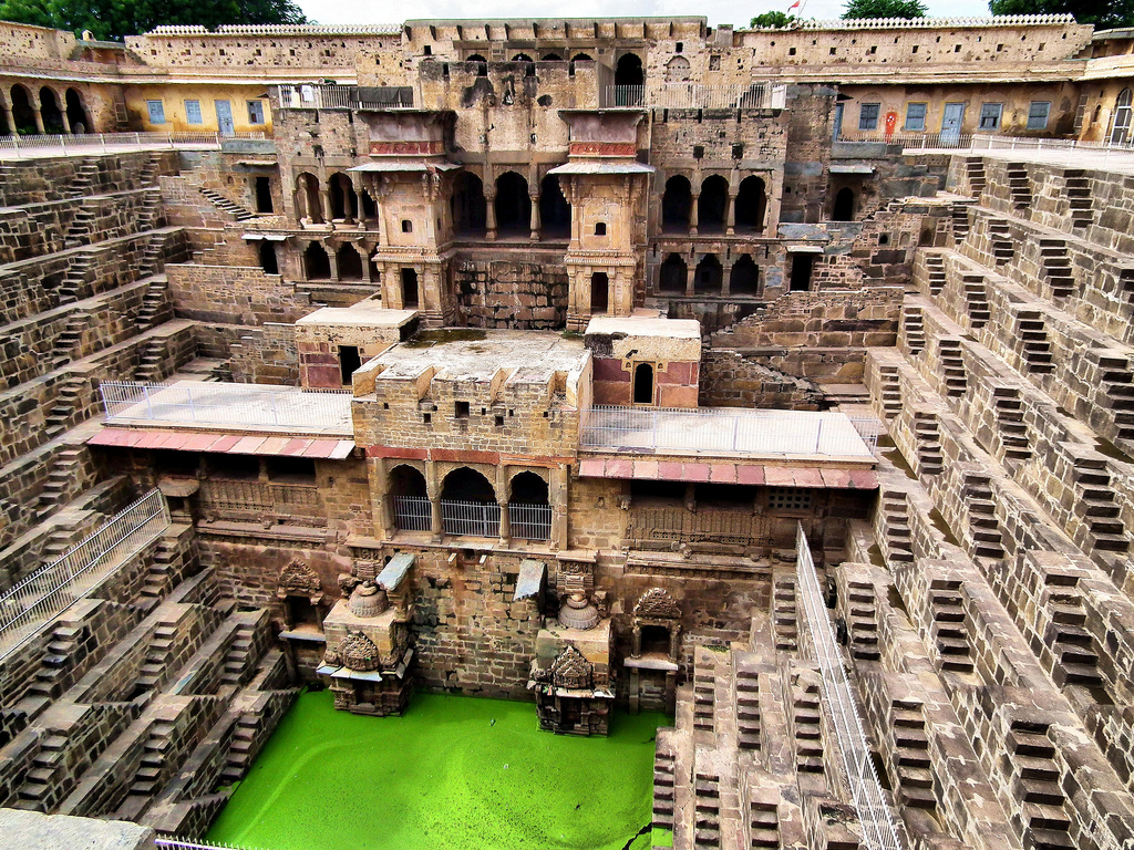 Deep stepped stone well with symmetrical geometric staircases descending to green algae-covered water, surrounded by ornate stone columns